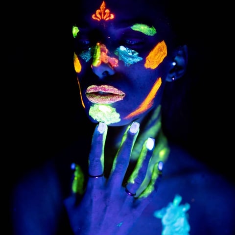 Person with glowing neon body paint and face art in vibrant colors against dark background