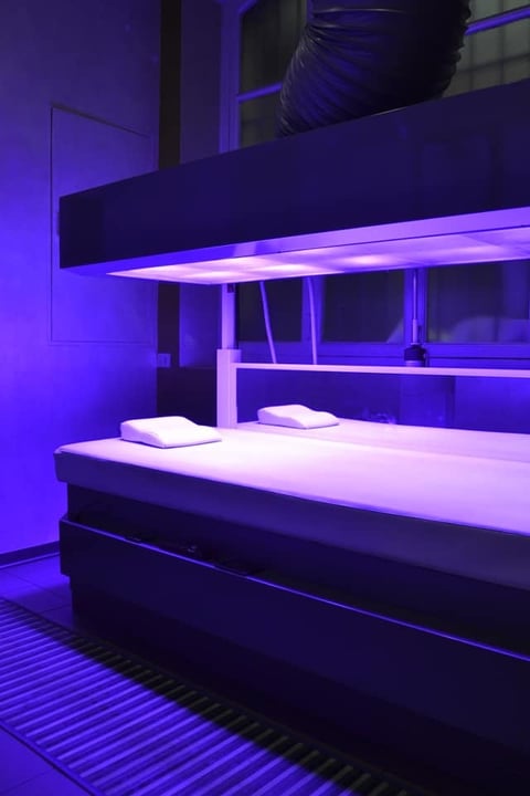 Modern bedroom with purple neon lighting, white bedding, sleek black furniture, and minimalist industrial design